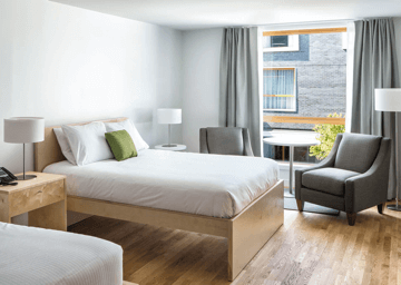 A daylit private room furnished with two full beds, bedside tables, and lamps, and by the open window two grey armchairs placed around a white circular table